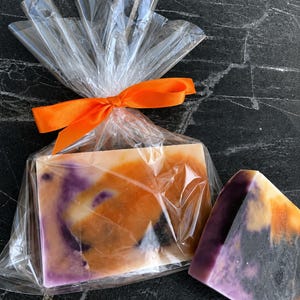 Op de afbeelding: Een rechthoekig stuk zeep met een gemarmerd patroon van oranje, paars en wit, verpakt in een doorzichtige plastic zak die is dichtgeknoopt met een oranje lint. Daarnaast ligt een tweede, kleiner driehoekig stuk van dezelfde zeep.