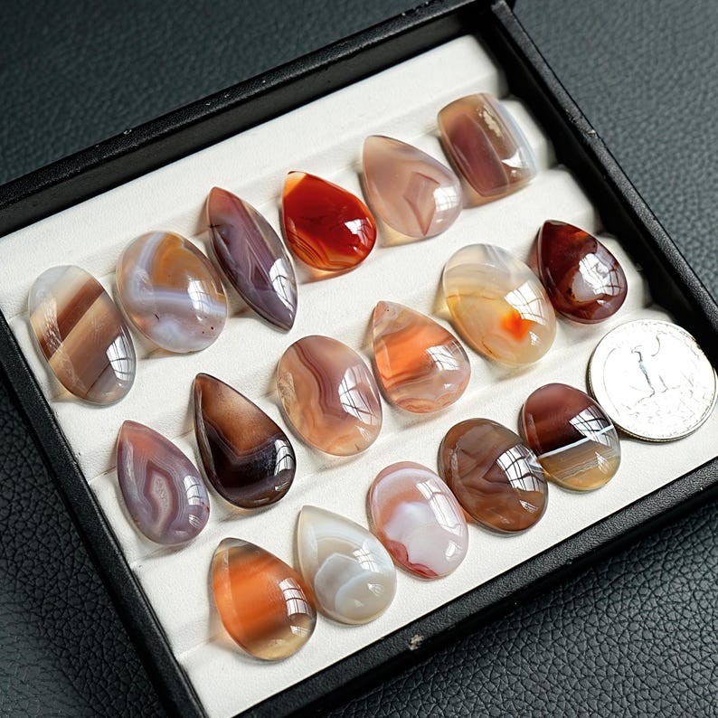 K&ouml;nnte beinhalten: Eine Sammlung polierter, mehrfarbiger Achat-Cabochons in ovalen und tropfenf&ouml;rmigen Formen, angeordnet in einer schwarzen Pr&auml;sentationsbox mit wei&szlig;en F&auml;chern. Die Steine weisen geb&auml;nderte Muster in Braun-, Orange-, Rot- und Wei&szlig;t&ouml;nen auf.