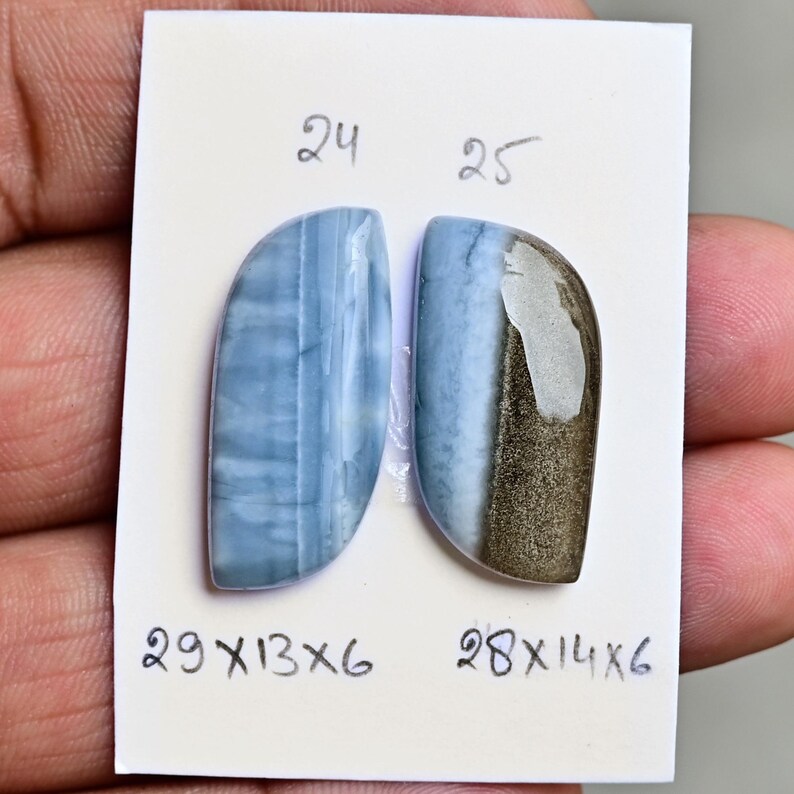 May include: Two polished, semi-circular blue gemstones mounted on a white card. The left stone is labeled 24 with dimensions 29x13x6. The right stone is labeled 25 with dimensions 28x14x6 and features a distinct metallic, bronze-colored section.