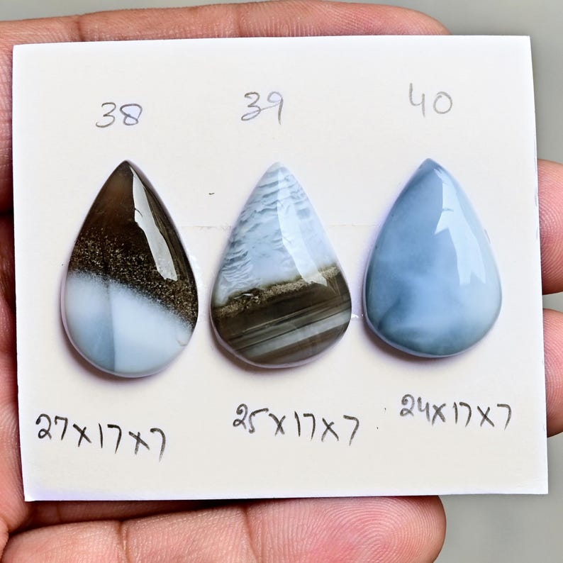 May include: Three pear-shaped cabochon gemstones arranged on a white card. The first, labeled 38, is brown and white with a measurement of 27x17x7 inches. The second, labeled 39, is blue and brown with a measurement of 25x17x7 inches. The third, labeled 40, is solid blue with a measurement of 24x17x7 inches.