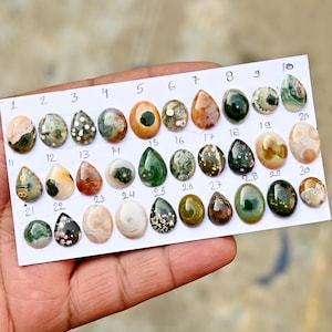 以下が含まれることがあります： 白いカードに並べられた、それぞれユニークな模様と色を持つ30個の磨かれた宝石のコレクション。 宝石は楕円形や涙滴形など様々な形をしており、緑、茶色、クリーム色の色合いを示しています。