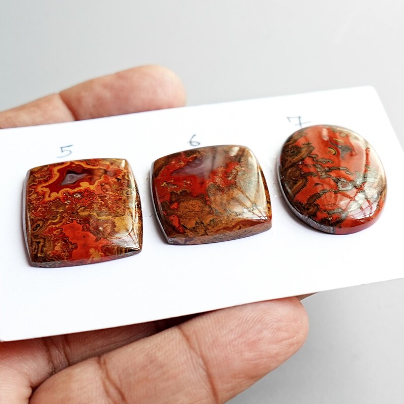 May include: Three polished cabochon gemstones arranged on a white card, labeled 5, 6, and 7. The stones feature intricate patterns of deep red, brown, and orange hues with swirling, agate-like inclusions and a smooth, reflective surface finish.