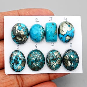 Pode incluir: Oito pedras preciosas de turquesa polidas dispostas em duas filas num cartão branco, numeradas de um a oito. As pedras apresentam tons de azul vibrantes com inclusões de pirite metálica, em várias formas ovais e retangulares com superfícies lisas.