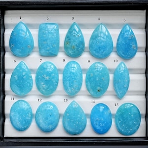 May include: A collection of fifteen polished, light blue gemstones in various shapes, including teardrops, ovals, and rectangles. The gemstones are displayed in a black tray with a white interior. Each stone has a unique pattern and slight variations in color.