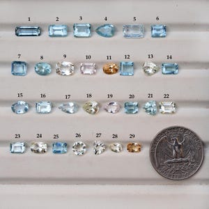 Peut inclure: Une collection de 29 pierres précieuses de formes et de couleurs variées, dont des teintes bleues, claires et dorées. Les pierres sont disposées sur une surface claire, avec une pièce de monnaie pour la comparaison des tailles.