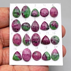 May include: A collection of twenty faceted ruby zoisite gemstones arranged on a white card, each labeled with a handwritten number from 1 to 20. The stones display a mix of vibrant magenta-pink and deep green colors in various shapes including ovals, pears, and triangles.