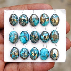 Puede incluir: Quince piedras preciosas de turquesa de forma ovalada con inclusiones de pirita metálica, dispuestas en tres filas sobre una tarjeta blanca. Cada piedra está numerada del 1 al 15 con tinta manuscrita, mostrando variados patrones azules y dorados.