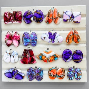 Può includere: Una collezione di sedici cabochon di pietre preziose lucide in varie forme e colori. Le pietre preziose includono sfumature di viola, arancione, blu, rosa e bianco, con motivi e texture unici. I cabochon sono disposti su una superficie bianca.