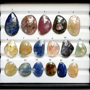 Peut inclure: Une collection de dix-sept pierres précieuses facettées de différentes formes et couleurs, dont le bleu, le rose, le jaune et le vert. Les pierres sont présentées dans un plateau noir avec des séparateurs blancs.