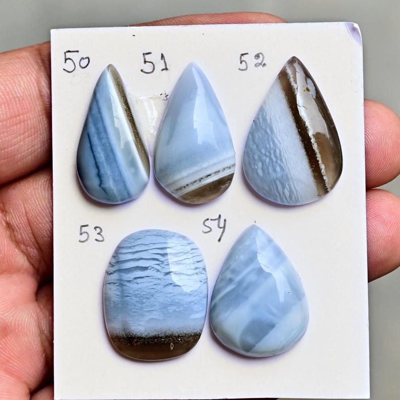 May include: Five polished blue and white banded gemstones arranged on a white card. The stones are labeled with the numbers 50, 51, 52, 53, and 54. The gems feature smooth, glossy surfaces with distinct horizontal and diagonal color patterns in shades of light blue, white, and brown.
