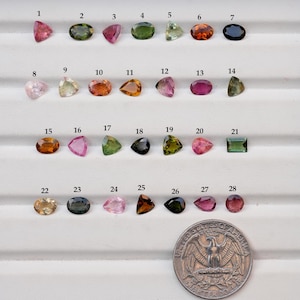 May include: A selection of 28 gemstones in diverse shapes and colours, including pink, green, orange, and blue. A quarter coin is included for size reference. The gemstones are arranged on a white surface.