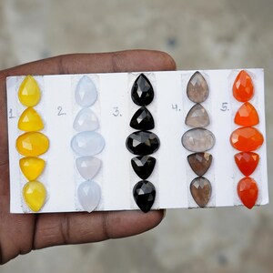 May include: A variety of gemstones in different shapes and colors are displayed on a white card. The gemstones include yellow, light blue, black, gray, and orange. Each color has a unique shape, such as teardrop, oval, and triangle.