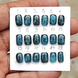 May include: Eighteen rectangular blue gemstones with dark inclusions, arranged in three rows on a white card. Each stone is numbered from 1 to 18 in handwritten ink above its position. The stones have a smooth, polished, cabochon-style surface.