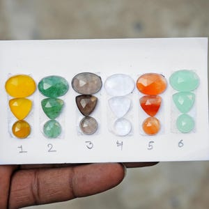 May include: Assortment of faceted gemstones in various shapes and colors, including yellow, green, gray, white, orange, and light green. The gemstones are arranged on a white card and numbered from 1 to 6.