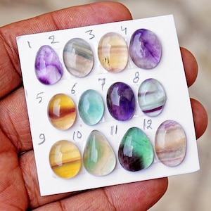May include: A collection of twelve polished, oval-shaped fluorite gemstones arranged on a white card. Each stone displays unique colour banding, including shades of purple, yellow, green, and clear white, with handwritten numbers one through twelve.