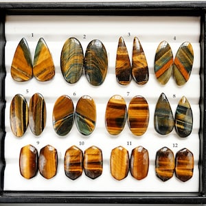 Puede incluir: Una colección de doce piedras preciosas de ojo de tigre pulidas en varias formas, incluyendo lágrima, ovalada y hexagonal. Las piedras muestran una mezcla de colores marrón dorado, azul y negro, con patrones y estrías naturales. Las piedras preciosas están dispuestas en una bandeja negra.