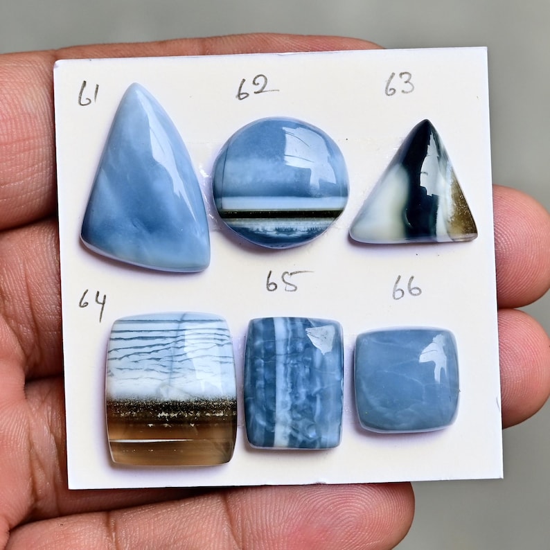 May include: Six polished cabochon gemstones arranged on a white card labeled 61 through 66. The stones feature various shades of blue, white, and translucent brown, with shapes including triangles, circles, and rectangles with smooth, reflective surfaces.