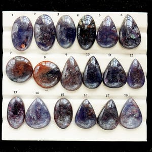 Peut inclure: Une collection de dix-huit pierres précieuses polies disposées en trois rangées sur un plateau de présentation blanc. Les pierres sont ovales, rondes et en forme de goutte, avec des teintes violettes, bleues et brun-rouge avec des inclusions.