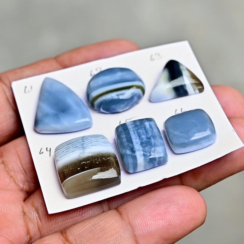 May include: Six polished cabochon gemstones arranged on a white card labeled with numbers 61 through 66. The stones feature various shades of blue, white, and brown with smooth, glossy surfaces in triangular, oval, square, and rectangular shapes.