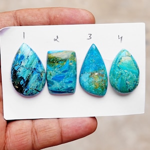 May include: Four polished gemstones arranged on a white card labeled 1 through 4. The stones feature shades of blue, teal, and green with dark veining and inclusions. Stone 1 is teardrop-shaped, 2 is rectangular, 3 is triangular, and 4 is oval.