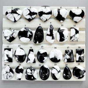Puede incluir: Una colección de veinticinco piedras preciosas blancas y negras en varias formas, incluyendo corazones, óvalos y rectángulos. Las piedras están dispuestas sobre una superficie blanca con etiquetas numeradas. Las piedras tienen un aspecto veteado.