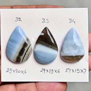 May include: Three pear-shaped cabochon gemstones arranged on a white card. Each stone is labeled with a number and dimensions in millimeters: 32 (29x20x6 mm), 33 (29x19x6 mm), and 34 (27x19x7 mm). The stones feature blue, white, and brown patterns.