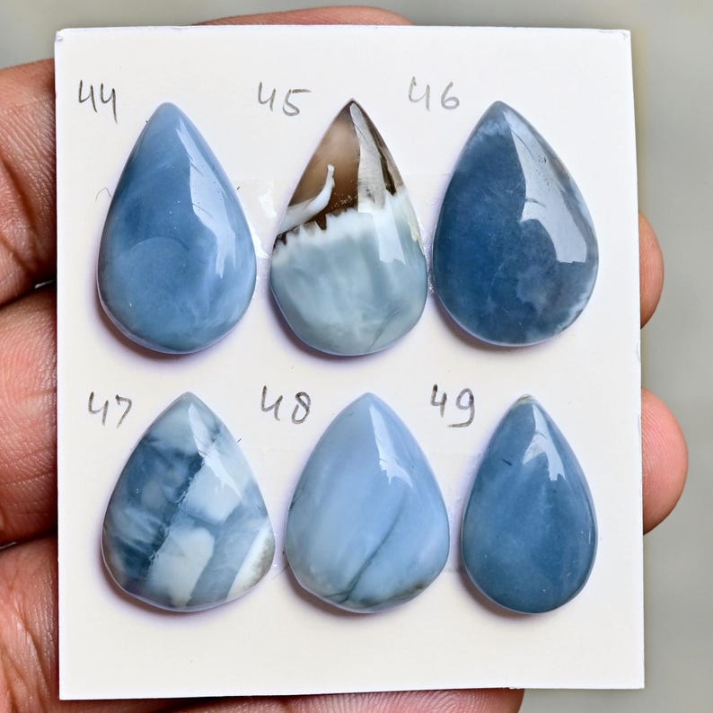 May include: Six polished, teardrop-shaped blue gemstones arranged in two rows on a white card. Each stone is labeled with a handwritten number from 44 to 49. The stones exhibit varying shades of blue, with one featuring a brown and white inclusion.