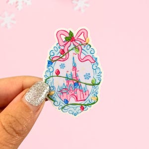 May include: A colorful sticker featuring a pink castle with a pink bow and Christmas lights. The sticker has a blue border and is held by a hand with a glittery nail. The background is pink with snowflakes and a pink ornament.