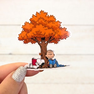 May include: A sticker featuring a cartoon scene of Snoopy and Charlie Brown under a tree with orange leaves. Snoopy is lying down reading a red book, while Charlie Brown sits reading a blue book. The tree has a brown trunk.