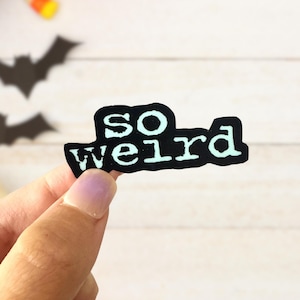 May include: A black sticker with the words "so weird" in a white, typewriter-style font. The sticker is held up against a white wooden background with Halloween decorations, including a pumpkin and bats.