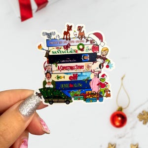 May include: A colorful sticker featuring a stack of Christmas movie titles and characters. Titles include "Home Alone," "Elf," and "Santa Clause." The sticker has a white border and is held by a hand with glitter nail polish.