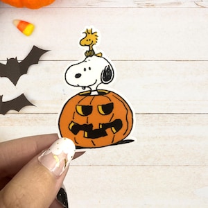 May include: A Halloween-themed sticker featuring Snoopy from Peanuts perched atop an orange jack-o'-lantern. The sticker has a white background and black outlines. Additional Halloween decorations, including candy corn and bats, are visible.