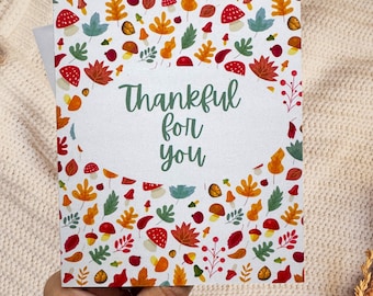 May include: A greeting card with a white background, featuring a colorful pattern of autumn leaves and mushrooms. The card has the text "Thankful for you" in green script. The card is held by a person.