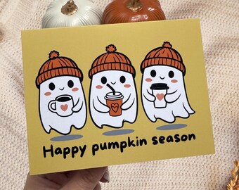 May include: A yellow greeting card with three cartoon ghosts wearing orange beanies and holding coffee cups. The card reads "Happy pumpkin season". Two decorative pumpkins are in the background.