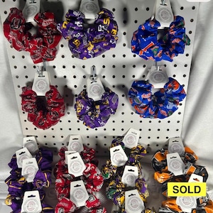May include: A collection of fabric hair scrunchies featuring various collegiate team logos and colors, including red, purple, blue, and orange patterns. Each scrunchie is attached to a white display card, with one item marked with a yellow SOLD label.