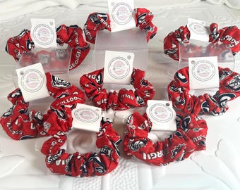 May include: A collection of seven red fabric hair scrunchies featuring a repeating pattern of bulldog graphics and the text Georgia Bulldogs. Each scrunchie is secured with a white paper tag that reads Handmade Items With Love.