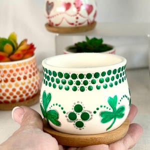 May include: A small, cream-colored ceramic planter featuring a hand-painted design of green shamrocks and metallic silver accents, topped with a band of green dots. The planter sits on a circular bamboo saucer, with other similar planters visible.
