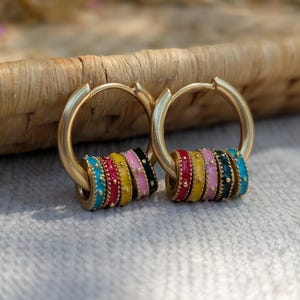 May include: Gold hoop earrings with colourful enamel beads. Each earring has a series of vibrant beads in turquoise, red, yellow, pink, and black. The earrings are displayed on a white surface with a woven background.