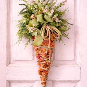 May include: A decorative, cone-shaped floral arrangement. The base is made of orange roses wrapped in twine. The top features greenery, pink flowers, and a green burlap bow. A gold ribbon is used for hanging. The arrangement is displayed against a white door.
