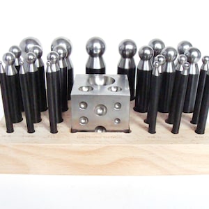 May include: A jewelry-making tool set featuring a wooden base holding various metal tools. The set includes a dapping block, metal punches with black handles, and ball-shaped tools. The tools are arranged neatly, ready for use in jewelry design and metalworking projects.