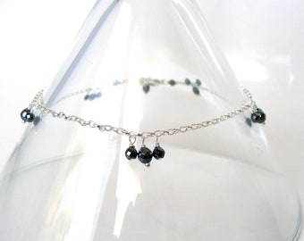 May include: A silver anklet with small, black faceted beads. The chain design features dangling clusters of three beads. The beads are a deep, reflective black, adding a touch of elegance. The anklet is a subtle piece of jewellery.