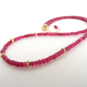 May include: A fine necklace composed of small, round, deep pink gemstones. Gold-coloured beads are placed at intervals along the necklace. The necklace is shown against a white backdrop.