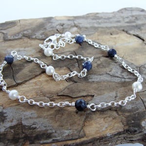 May include: A delicate silver chain necklace featuring alternating white pearls and faceted dark blue gemstones. The necklace is displayed on a textured, brown wooden surface, showcasing its elegant design.