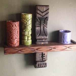 May include: A wooden shelf with four decorative tiki-themed items. The shelf holds a brown, green, and black tiki statue, and a small purple cup. The shelf is made of wood and has a carved design.