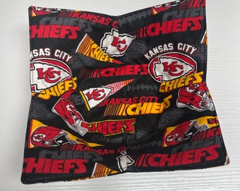 May include: A black and red fabric bowl cozy featuring the Kansas City Chiefs logo and team name. The design includes the team's arrowhead logo, helmets, and the words "Chiefs" and "Kansas City". The cozy is square-shaped.