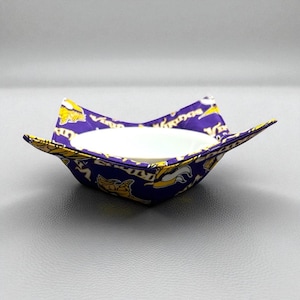 Puede incluir: Un acogedor cuenco de tela morada y dorada con un cuenco de cerámica blanco en el interior. La tela presenta un patrón repetido del logotipo de los Minnesota Vikings. El acogedor está diseñado para sostener un cuenco y mantenerlo caliente.