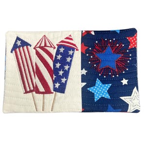 May include: A quilted fabric item featuring patriotic designs. One side displays embroidered red, white, and blue fireworks. The other side has a navy blue background with white stars and red and blue firework bursts. The item is likely a small pouch or wallet.