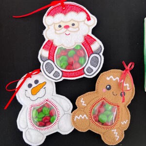 May include: Three Christmas-themed felt pouches: Santa Claus, snowman, and gingerbread man. Each has a clear window filled with red and green candies, and a red ribbon for hanging. The Santa pouch is red and white, the snowman is white, and the gingerbread man is brown.