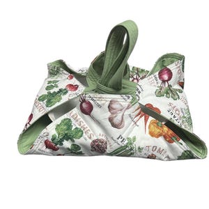 May include: A white and green quilted casserole carrier with a handle. The carrier features a vegetable print with illustrations of radishes, garlic, carrots, and artichokes. The carrier is designed to hold a dish and has a green handle.