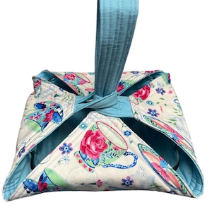 May include: A quilted bowl cozy with a light blue handle. The cozy features a floral pattern with teacups, roses, and butterflies on a cream background. The interior and trim are light blue.